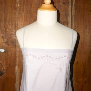 May include: A light lavender slip dress featuring thin spaghetti straps and a straight neckline. The upper chest area is decorated with delicate, tonal floral embroidery that includes small stitched roses and hanging linear details.