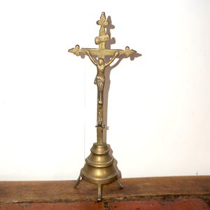 Puede incluir: Crucifijo de latón con una figura detallada de Jesús en la cruz. La cruz tiene extremos decorativos y se asienta sobre una base escalonada con tres pequeños pies. La pieza tiene una pátina cálida y envejecida.