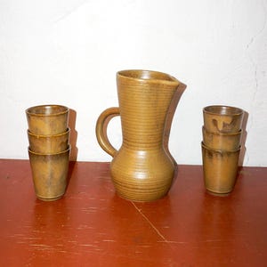Op de afbeelding: Een keramische kan met een handvat en tuit, vergezeld van zes bijpassende kleine bekers. De set heeft een warme, aardse bruine kleur met een gestructureerd, geribbeld ontwerp. De kan is ongeveer 25 cm hoog.