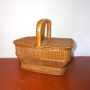 Könnte beinhalten: Ein geflochtener Picknickkorb mit einem Griff. Der Korb ist rechteckig mit einem gebogenen Boden und einem Deckel. Der Griff ist gewölbt und aus dem gleichen geflochtenen Material gefertigt. Der Korb hat eine hellbraune Farbe.
