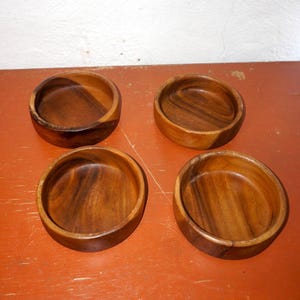 Könnte beinhalten: Vier kleine, runde Holzschalen in einem satten, dunklen Braunton. Die Schalen haben eine glatte, polierte Oberfläche und sind quadratisch angeordnet. Die sichtbare Holzmaserung unterstreicht ihr natürliches Aussehen. Jede Schale hat einen Durchmesser von ca. 10 cm.
