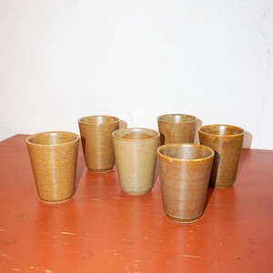 May include: Six brown ceramic mugs on a red wooden surface. The mugs are all the same size and shape, and they have a slightly textured finish.