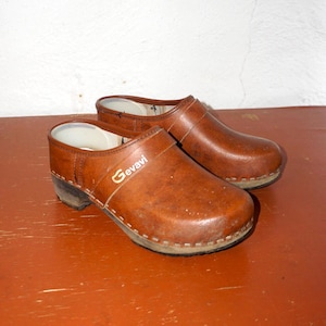 Può includere: Un paio di zoccoli in pelle marrone con suola in legno. Gli zoccoli hanno la punta arrotondata e una cinghia sulla parte superiore. Il marchio "Gevavi" è visibile sul lato. Sono su una superficie marrone-rossastra. Stile classico.