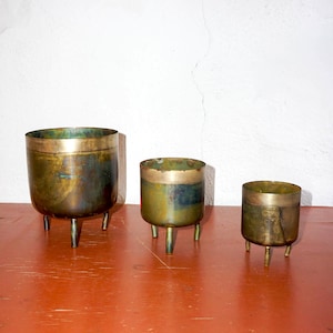 May include: Three gold metal planters with a distressed finish and three legs each. The planters are arranged in order of size from left to right.