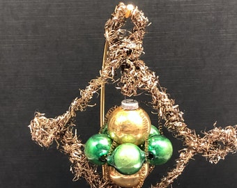 Puede incluir: Adorno navideño decorativo con un grupo de bolas verdes y doradas. Las bolas están rodeadas por un marco de alambre dorado y material similar a oropel. El adorno está sobre un soporte de alambre dorado sobre un fondo negro.