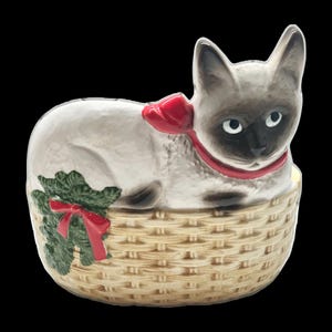 May include: A ceramic Siamese cat figurine in a woven basket. The cat has a red ribbon around its neck and a red bow with green foliage on the basket. The cat has a white body, dark brown face, ears, and paws.