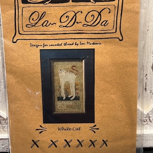 May include: A craft kit for counted thread embroidery. The packaging is tan with black text and a framed image of a white cat. The text reads "La-D-Da" and "White Cat". The kit includes designs by Lori Markovic.