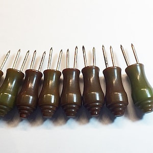 May include: A set of nine corn cob holders with smooth, olive-green, hourglass-shaped handles and two sharp, silver-toned metal prongs extending from the top of each piece. The handles feature a tiered, circular base design at the bottom.