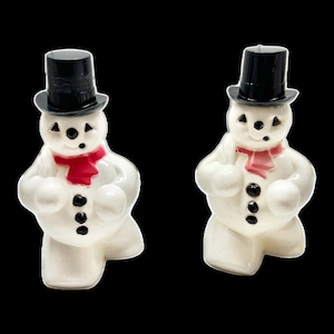 May include: Two white snowman figurines with black top hats and coal button eyes and mouths. Each snowman wears a red scarf. The snowmen are standing on a black background.