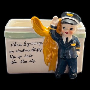 Puede incluir: Una maceta de cerámica vintage con una figura de joven piloto con uniforme azul marino, saludando. La maceta tiene una base blanca con una franja verde claro y el texto "When I grow up, an airplane I'll fly, Up, up into the blue sky."