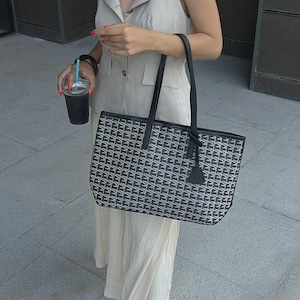 Könnte beinhalten: Eine schwarz-weiß gemusterte Tragetasche mit schwarzen Griffen und einem schwarzen Anhänger. Die Tasche ist rechteckig und hat ein geometrisches Design. Die Person trägt ein beiges Kleid und hält ein Getränk mit einem blauen Strohhalm.