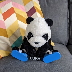 May include: A black and white panda plush toy wearing blue and white leg braces with the word "LUKA" printed on the front. The panda is sitting on a gray couch with a colorful patterned pillow behind it.