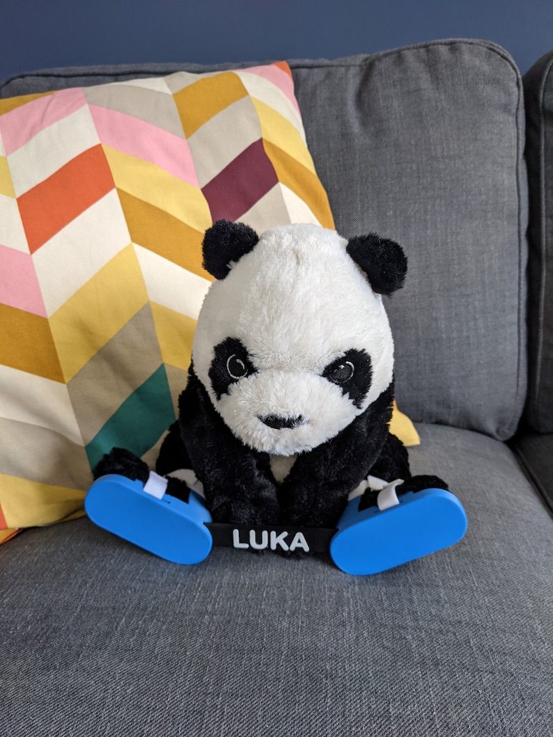 May include: A black and white panda plush toy wearing blue and white leg braces with the word "LUKA" printed on the front. The panda is sitting on a gray couch with a colorful patterned pillow behind it.