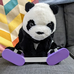 May include: A black and white panda plush toy wearing purple slippers. The panda is sitting on a white surface with a colourful patterned pillow behind it.