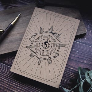May include: A brown notebook with a black geometric design featuring weapons and a central emblem. A dark wood fountain pen with a gold-colored nib and cap rests on a wooden cutting board. The scene is set on a dark wood surface.