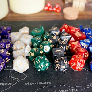 May include: A collection of colourful dice with gold numbers, including purple, white, green, black, red, and blue dice. The dice are on a black surface with a map pattern.
