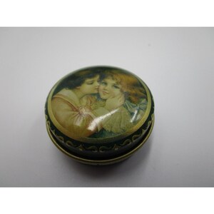 May include: A round, green and gold metal tin with a picture of two children on the lid. The children are embracing and have a sweet expression on their faces.