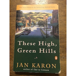 May include: Book cover for "These High, Green Hills" by Jan Karon, the third novel in the bestselling Mitford series. The cover features a painting of a quaint town street with a car and a bicycle.