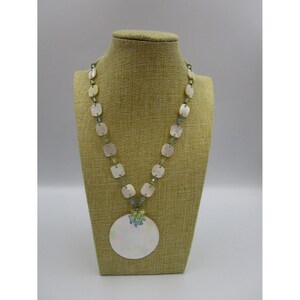 May include: A white and green shell necklace with a large, round shell pendant. The necklace is on a fabric-covered mannequin.