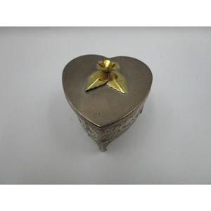 May include: A silver heart-shaped trinket box with an ornate design and three legs. The lid features a gold flower with leaves.