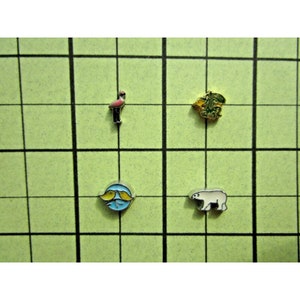 Puede incluir: Cuatro pequeños encantos sobre un fondo de cuadrícula verde. Los encantos son un flamenco rosa, una planta de hojas verdes y doradas, un círculo azul con dos peces amarillos y un oso polar blanco.