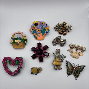 Puede incluir: Una colección de diez broches decorativos variados que incluyen una cesta de flores, una planta en maceta, bellotas, un coche antiguo, un broche floral morado oscuro, un conejo, una corona de rosas en forma de corazón, una oveja y una mariposa.