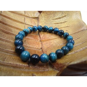 May include: A blue tiger eye bead bracelet. The bracelet features polished, round beads in varying shades of blue and black. The beads are strung together on an elastic band. The bracelet is displayed on a wooden surface.