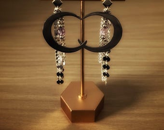 May include: A pair of black crescent moon-shaped earrings hanging from a gold-toned T-bar display stand. Each earring features a dangling silver-toned chain with small metallic discs and a single light purple bead accent attached to the black moon.