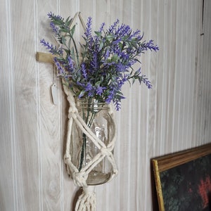 May include: A glass jar with a bouquet of purple lavender flowers is hanging from a macrame plant hanger with a white tassel. The hanger is attached to a wooden dowel.