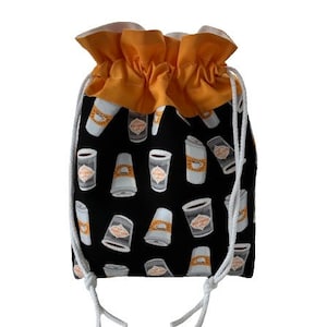 May include: A black drawstring bag with a repeating pattern of coffee cups in white and tan. The top of the bag is a ruffled orange fabric, and the drawstring is white.