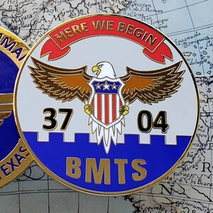 May include: Two gold-rimmed military challenge coins. One coin features a torch and wings with text "AIR TRAINING COMMAND LACKLAND AFB TEXAS". The other coin has an eagle, shield, and text "HERE WE BEGIN 37 04 BMTS".