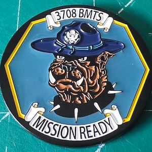 May include: A round, black and yellow patch with a bulldog wearing a blue hat. The patch has the text "3708 BMTS" and "MISSION READY". The bulldog is brown and white, with a blue background.