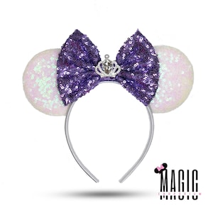 Puede incluir: Una diadema con orejas de ratón con lentejuelas blancas y un gran lazo con lentejuelas moradas. Una pequeña corona plateada está centrada en el lazo. La diadema es plateada. El logotipo de Magic Earporium está en la esquina inferior derecha.