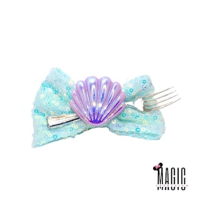 May include: A light blue sequined bow with a silver fork and a pearlescent seashell accent. The bow is decorated with small, shiny sequins. The seashell is iridescent, reflecting shades of purple and blue. The bow is a hair accessory.