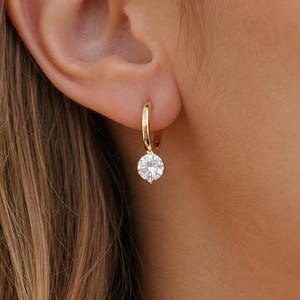 May include: A gold hoop earring featuring a single round, clear, faceted gemstone charm suspended from the bottom. The metal has a polished, reflective finish, and the stone is secured in a simple prong setting, catching the light clearly.