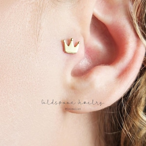 Puede incluir: Un pendiente de oro en forma de corona, usado en el cartílago superior de la oreja. La corona es un diseño simple con una parte superior redondeada y cinco puntas.
