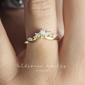 Può includere: Un anello dorato con un design a corona, con più pietre preziose trasparenti. L'anello è indossato su un dito. L'immagine include il nome del marchio "Goldspoon Jewelry" e la parola "Minimalist".