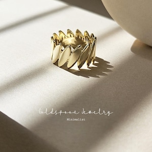 May include: A gold ring with a minimalist design featuring a series of overlapping oval shapes. The ring is photographed on a light beige surface.