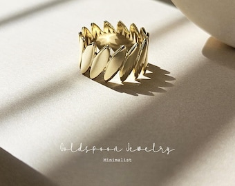 Può includere: Un anello in oro con un design minimalista che presenta una serie di forme ovali sovrapposte. L'anello è fotografato su una superficie beige chiaro.