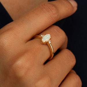 May include: A gold ring with a white opal gemstone and two smaller diamonds on the band. The ring is on a person's finger.