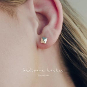 May include: A close-up of a gold-colored, geometric stud earring on an ear. The earring has a pyramid shape with a flat, reflective surface. The text "Goldspoon Jewelry" and "Minimalist" is visible.