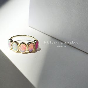 May include: A gold ring featuring six oval-shaped opals in shades of pink, white, and purple. The ring is set against a white background with a shadow. The text "Goldspoon Jewelry Minimalist" is in the background.