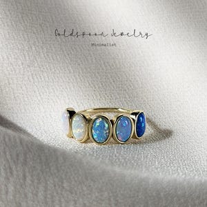 May include: A gold ring featuring five oval-shaped opals in a row, transitioning in color from white to deep blue. The text Goldspoon Jewelry Minimalist is visible above the ring, which is set against a textured, off-white fabric background.