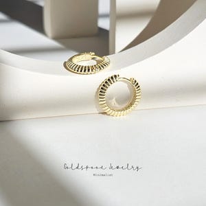 Puede incluir: Dos pendientes de aro dorados con un diseño texturizado y acanalado. Los pendientes están sobre una superficie blanca. Goldspoon Jewelry Minimalist.