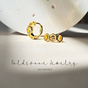 May include: A pair of gold hoop earrings with a honeycomb design. The earrings are on a white surface with the text "Goldspoon Jewelry California" below them.