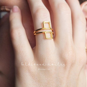 May include: Gold geometric ring with two stacked square shapes. The ring is worn on a person's finger.