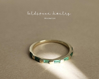 May include: A gold band ring with a row of emerald green rectangular stones. The ring is on a light brown surface. Goldspoon Jewelry Minimalist.
