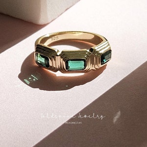 Puede incluir: Anillo de oro con tres piedras preciosas verdes engastadas en un diseño geométrico. El anillo está marcado con "925" en la banda interior.