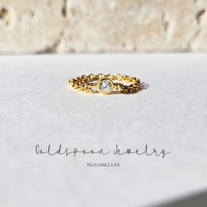 Op de afbeelding: Een gouden kettingring met een enkele ronde, heldere steen in het midden. De ring ligt op een wit oppervlak met de tekst "Goldspoon Jewelry Minimalist" eronder.