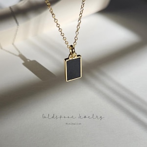 Puede incluir: Un collar de cadena dorada con un colgante rectangular negro. El colgante tiene un borde dorado. El collar está sobre una superficie blanca. Goldspoon Jewelry Minimalist.